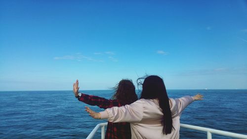 Rear view of friends with arms outstretched standing in boat on sea against blue sky