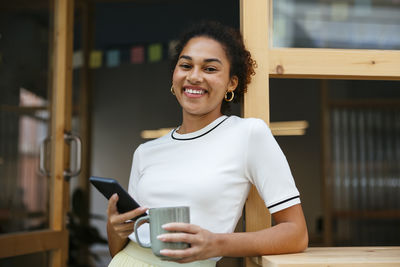 Smiling student standing with coffee cup and smart phone in doorway