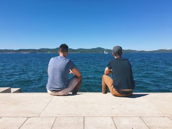 Rear view of friends sitting by river against clear blue sky