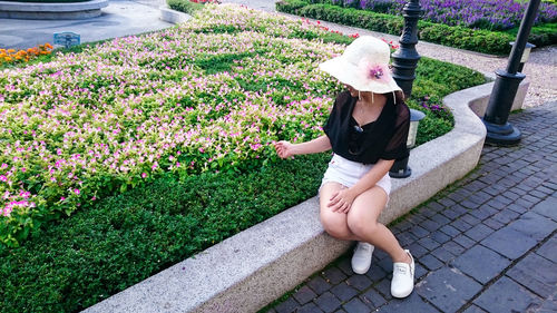 Full length of woman holding flowering plants while sitting on retaining wall in city