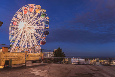 Illuminated ferris wheel in city against blue sky