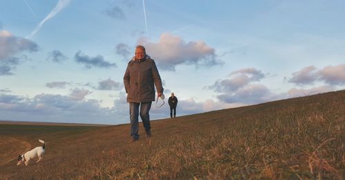 Mature woman and man with dog on grassy field against sky