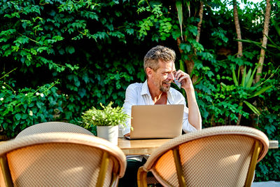 Happy mature male manager in white shirt with fresh drink and speaking on smartphone while sitting at table with laptop during work in street restaurant in summer