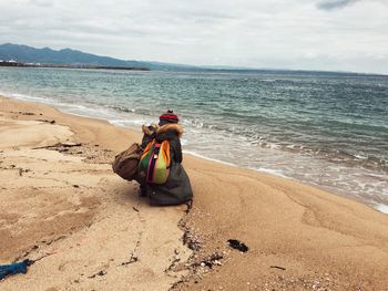 Rear view of woman with backpacks crouching at beach against sky