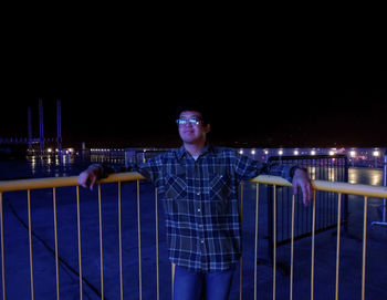 Portrait of young man standing against railing at night