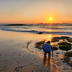 Rear view of boy crouching at beach during sunset