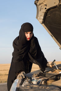 Portrait of woman repairing car while standing on road against sky