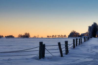 Wooden fence on snow covered field against sky during sunset