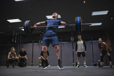 Adaptive athlete jumping while lifting barbell with people cheering in background at gym