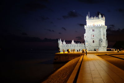 People at illuminated building by sea against sky at night