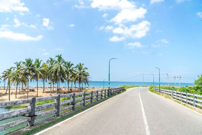 Empty road by palm trees against sky