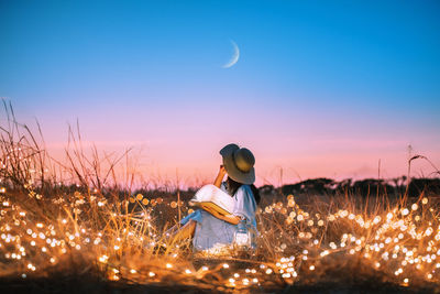 Young woman sitting with book in an illuminated field against sky