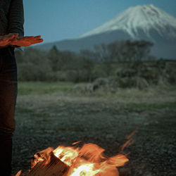 Midsection of man standing by bonfire warming hands  with  mountain background.