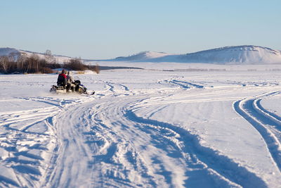 The snowmobile with fishermen is riding along the winter lake against the backdrop of the mountains. 