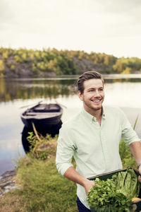 Smiling man carrying vegetables crate while standing at lakeshore