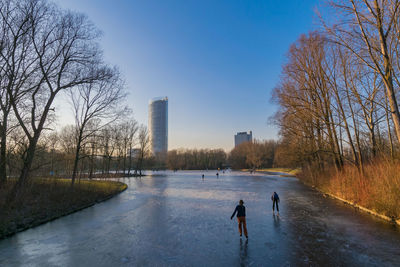 Man standing by frozen river in city against clear sky during winter