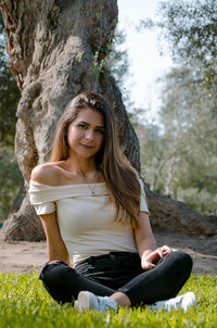 Full length portrait of smiling young woman sitting against trees in park