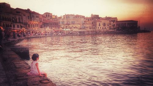 Woman sitting on canal in city against sky at sunset