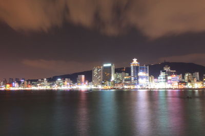 Illuminated buildings by sea against sky at night