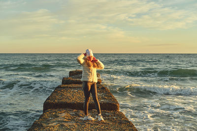 A woman with blond hair in a hat and a fur coat walks along the sea along a stone breakwater