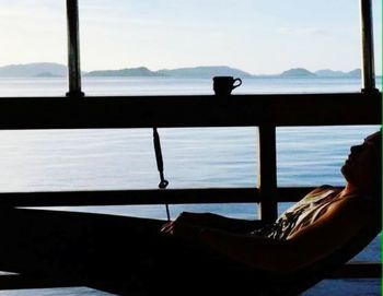 Silhouette man relaxing by window against sea