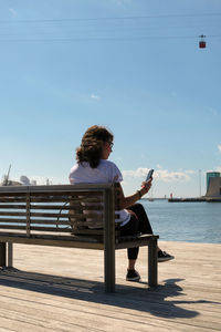 Woman using mobile phone while sitting on bench against sea