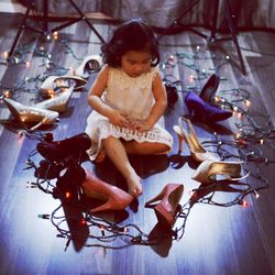 Girl sitting amidst high heels and christmas lights on hardwood floor