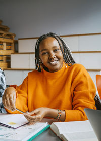 Portrait of smiling young female student with braided hair sitting at desk in college classroom