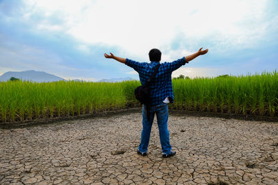 Rear view of man with arms outstretched standing on dry field against sky