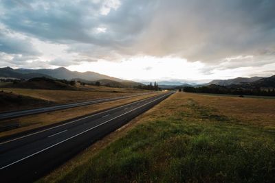 Road passing through field against cloudy sky