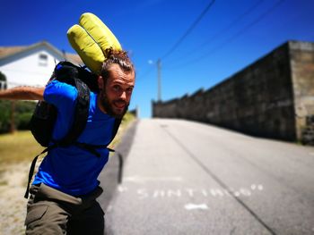 Portrait of young man with backpack standing on road against clear blue sky during sunny day