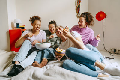 Cheerful female friends watching tv while enjoying snacks at home