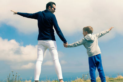 Back view of happy father and son holding hands and having fun against the sky at mountain peak.