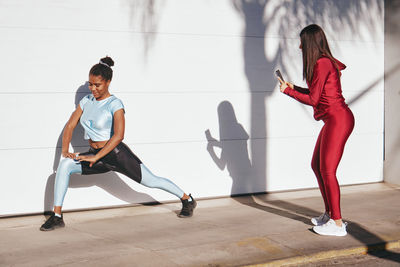 Full body of young athletic female in stylish sportswear taking photo of positive african american sportswoman doing lunges exercise against white wall on street