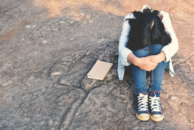 High angle view of woman hugging knees while sitting by book on rock