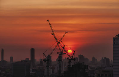 Silhouette cranes by buildings against sky during sunset