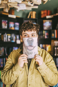 Playful young man with curly hair covering face with book in bookstore