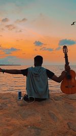 Rear view of man sitting on beach against sky during sunset