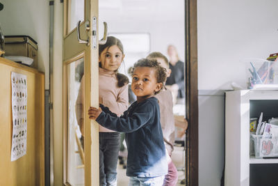 Boy opening door while standing with classmates at doorway of kindergarten