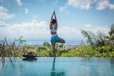 Rear view of young woman with arms raised standing by infinity pool