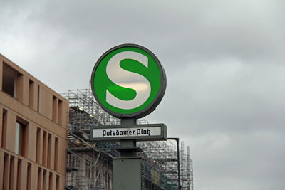 Low angle view of underground subway sign by building against cloudy sky