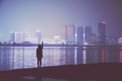 Silhouette woman doing peace sign by lake against buildings at night