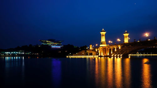 Illuminated buildings by river against blue sky at night