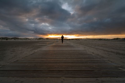 Rear view of woman standing on boardwalk at sandy beach against sky during sunset