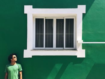 Young man looking away while standing by closed window on green wall