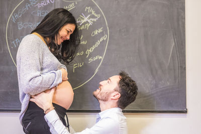 Side view of man kneeling in front of pregnant girlfriend against blackboard