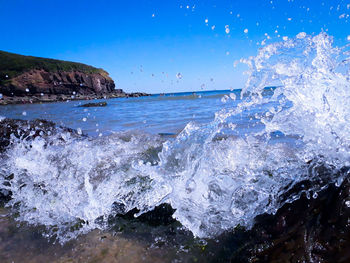 Waves splashing on rocks at shore against blue sky