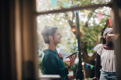 Happy young woman talking through earphones while standing with friend in balcony during party