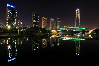 Illuminated modern buildings by river against sky at night