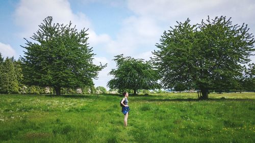 Side view of young woman standing on grassy field against cloudy sky